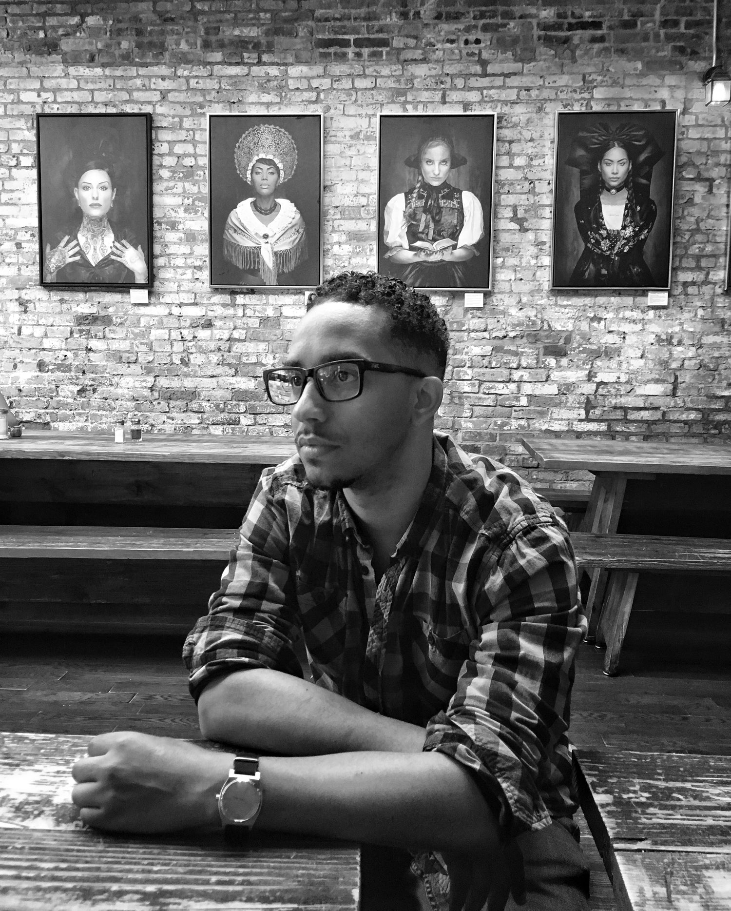A black and white photo of the artist Corey Jones in a rustic restaurant with custom portrait paintings in the background 